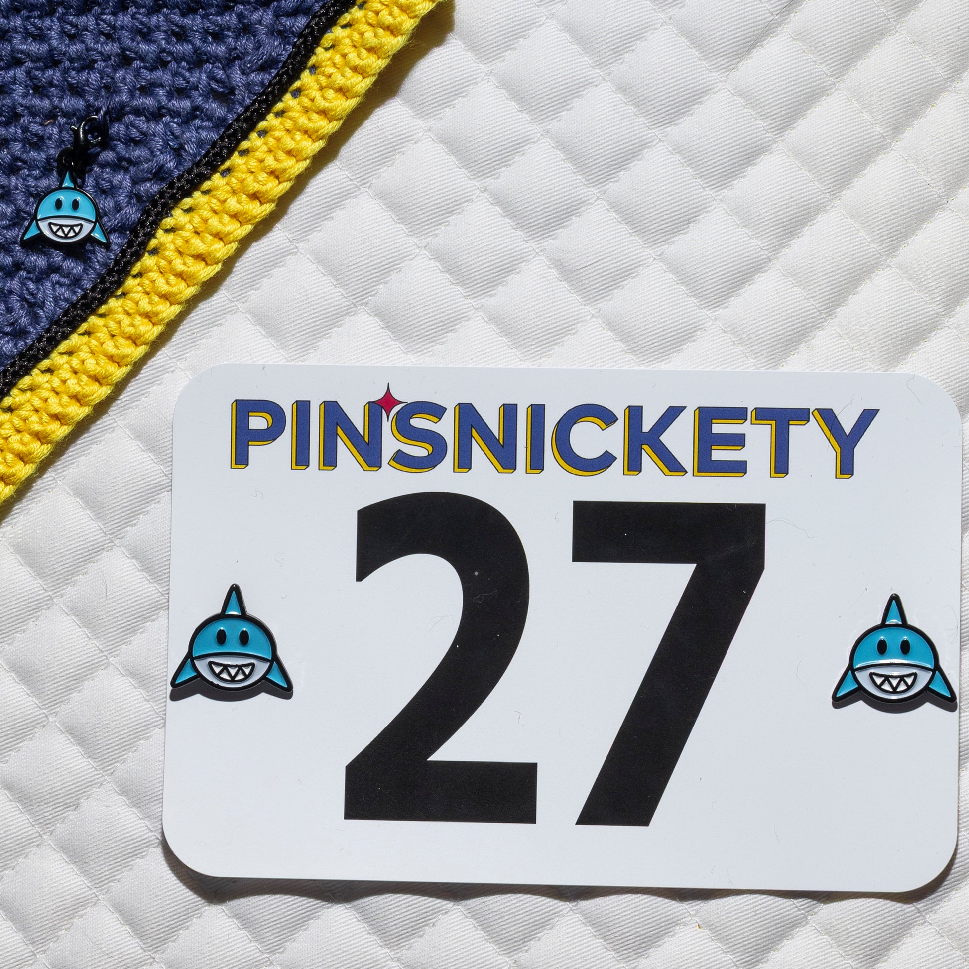 a pinsnickety shark bridle charm clipped to a purple jumper bonnet beside a pair of shark horse show number pins holding a show number onto a white saddle pad.