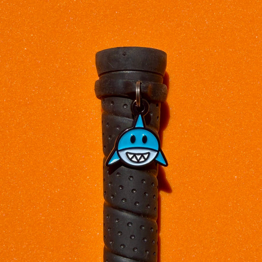 A black textured horseback riding whip handle with a blue and white pinsnickety shark crop charm attached near the top, set against a solid orange background.