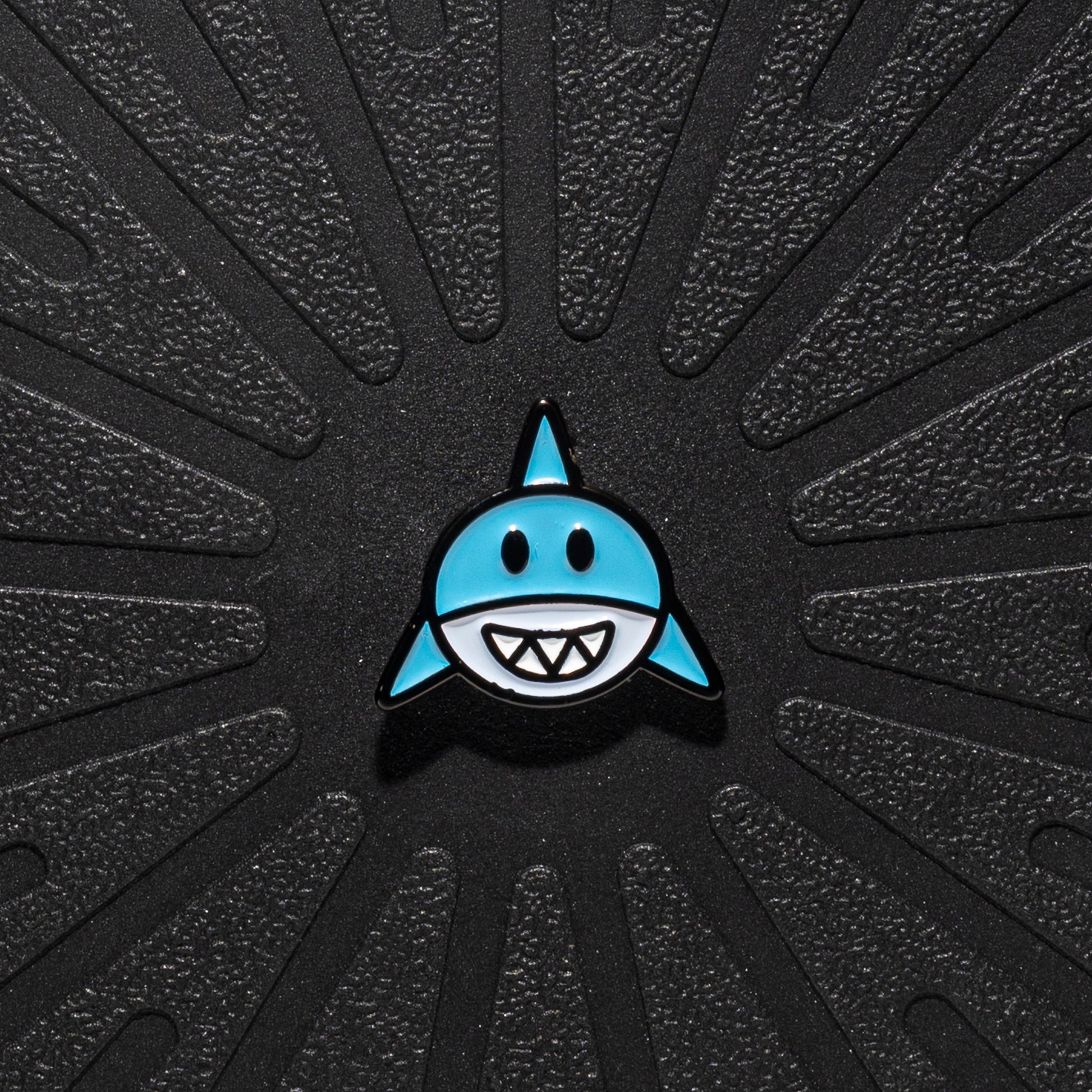 A single Pinsnickety horse show number pin shaped like a shark with a light blue body, black outline, and white teeth, centered on a textured black background with radiating grooves.