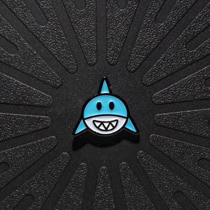 A single Pinsnickety horse show number pin shaped like a shark with a light blue body, black outline, and white teeth, centered on a textured black background with radiating grooves.
