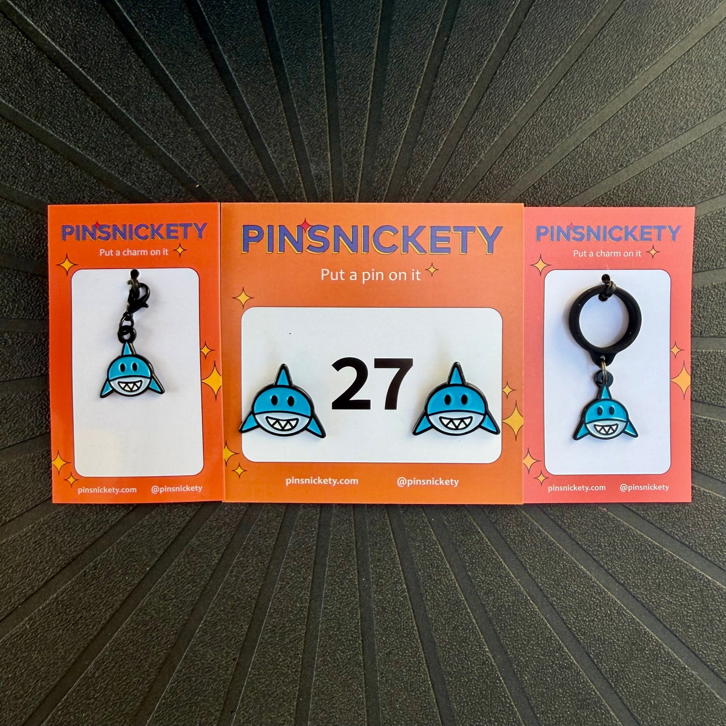 Three orange PINSNICKETY cards displaying shark-themed horse show number pins, a bridle charm, and a crop charm, arranged on a black textured background. The center card shows two blue and white shark pins with the number 27, while the left and right cards show shark charms.