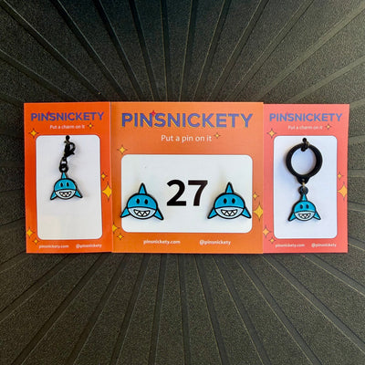 Three orange PINSNICKETY cards displaying shark-themed horse show number pins, a bridle charm, and a crop charm, arranged on a black textured background. The center card shows two blue and white shark pins with the number 27, while the left and right cards show shark charms.