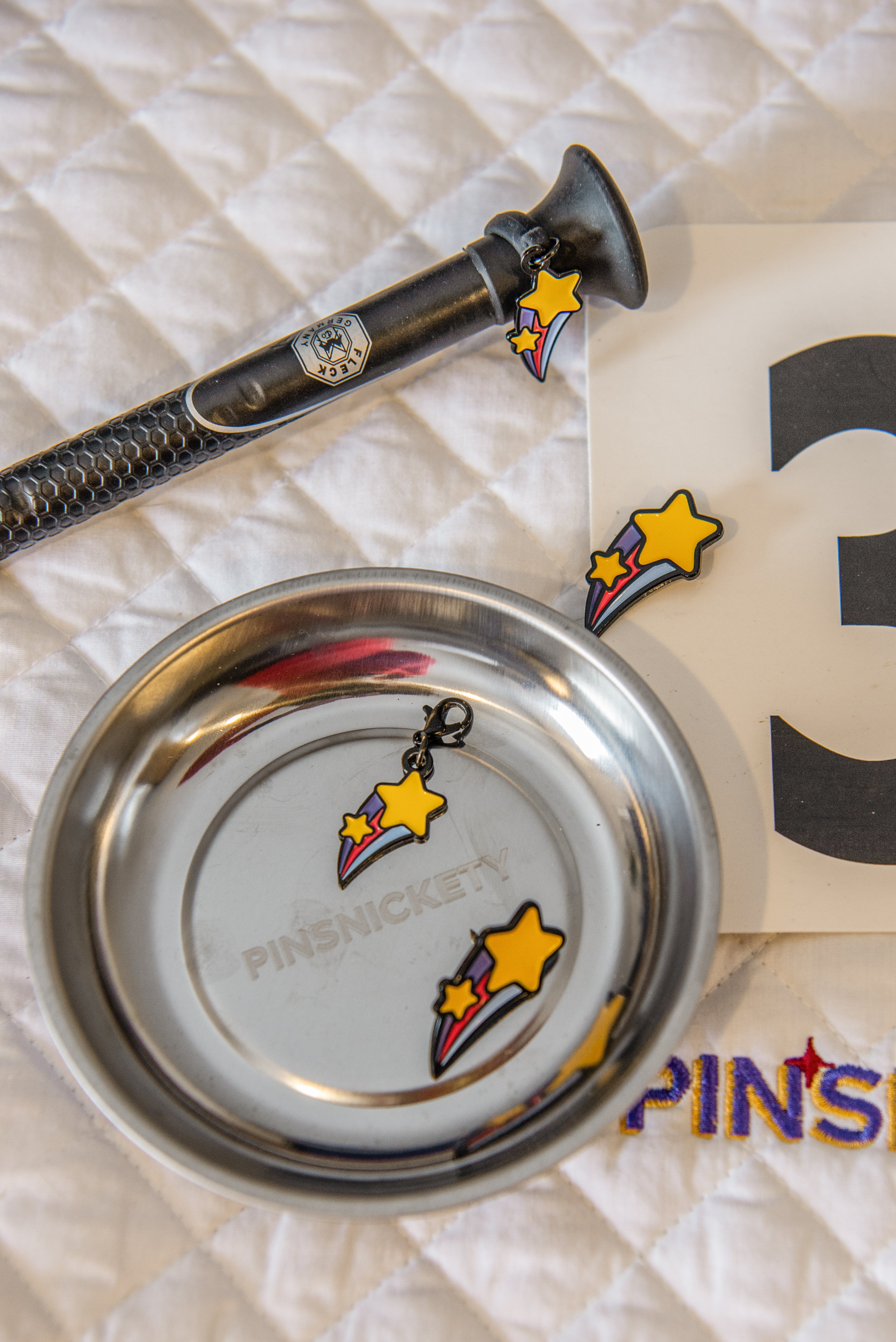 a white saddle pad with a a crop, show number, and magnetic storage dish all decorate wtih pinsnickety shooting star number pins and charms.