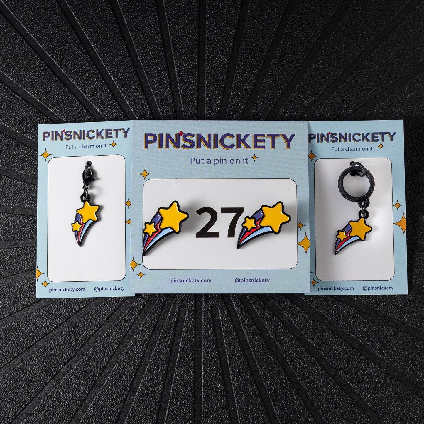 A matching triple combination set with Pinsnickety Shooting Star horse show number pins, a bridle charm, and a crop charm in their product packaging on a black background.