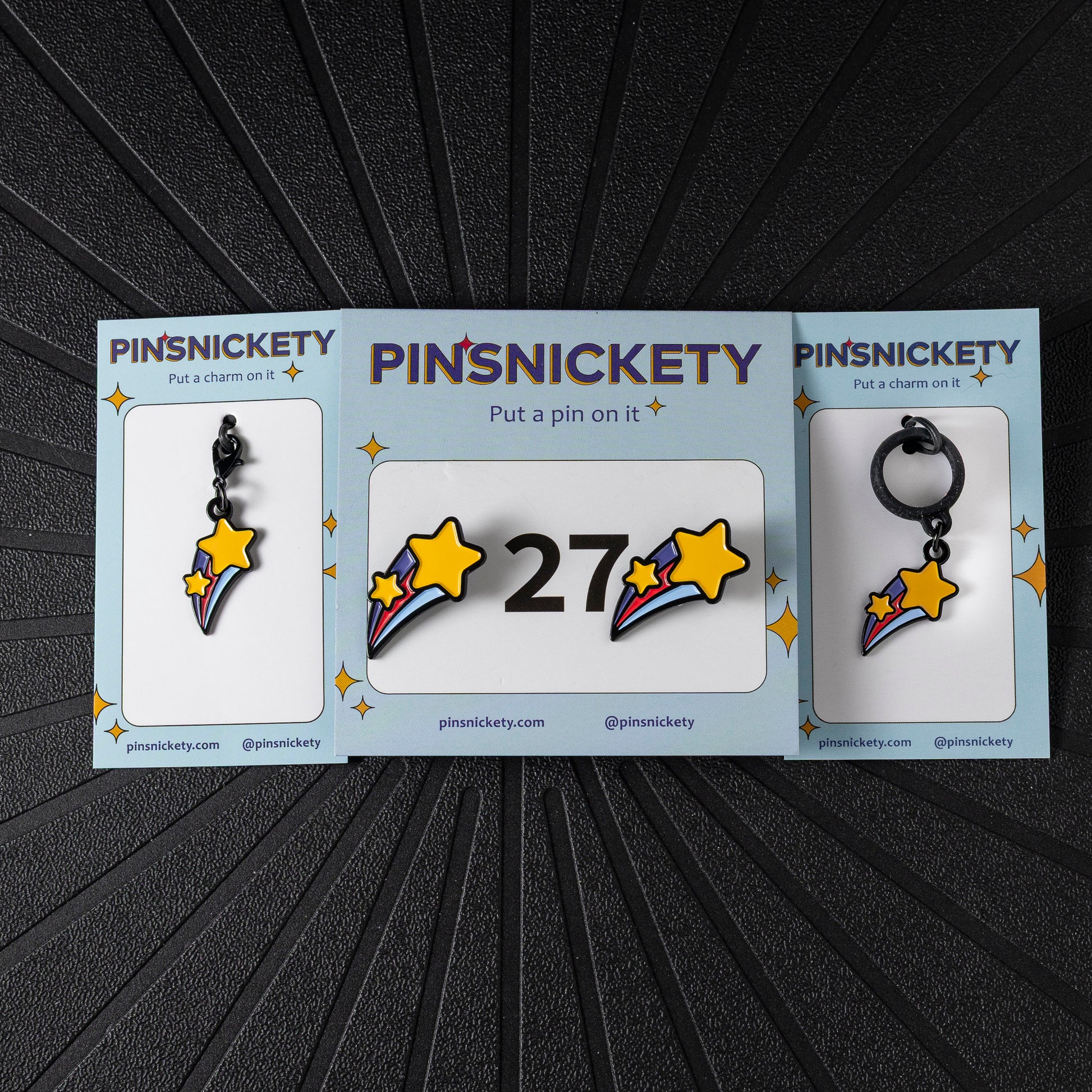 A matching triple combination set with Pinsnickety Shooting Star horse show number pins, a bridle charm, and a crop charm in their product packaging on a black background.