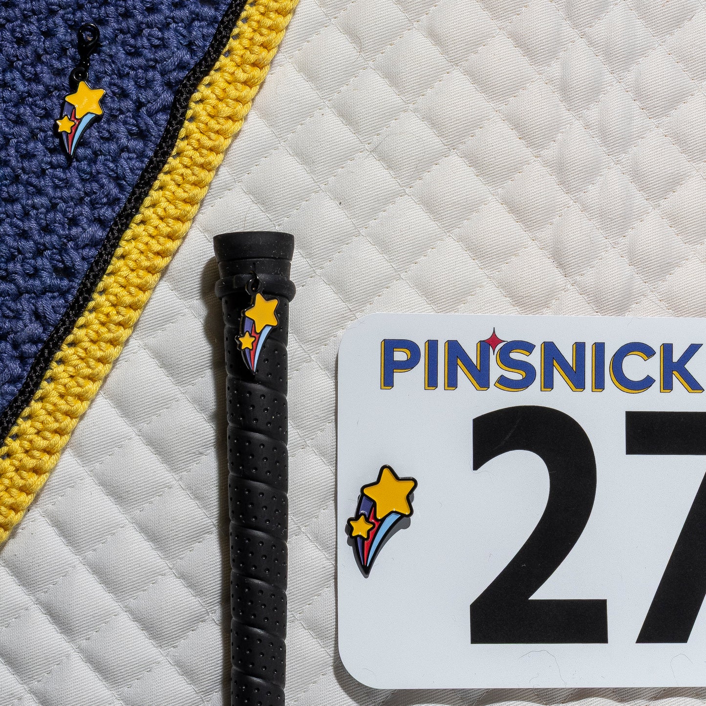 A saddle pad, bonnet and whip decorated with matching Shooting Star Pinsnickety horse show number pins, a bridle charm and a crop charm.