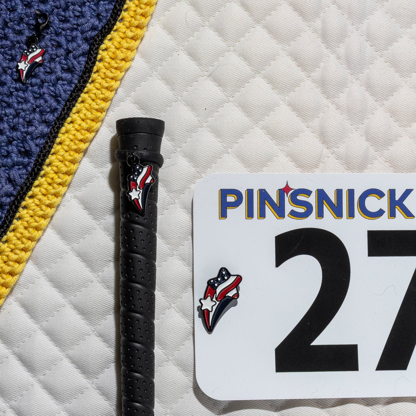A saddle pad, bonnet and whip decorated with matching Stars and stripes Pinsnickety horse show number pins, a bridle charm and a crop charm.