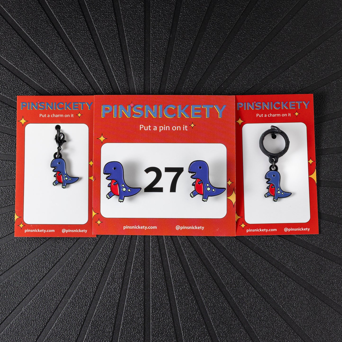A matching triple combination set with Pinsnickety T-Rex horse show number pins, a bridle charm, and a crop charm in their product packaging on a black background.