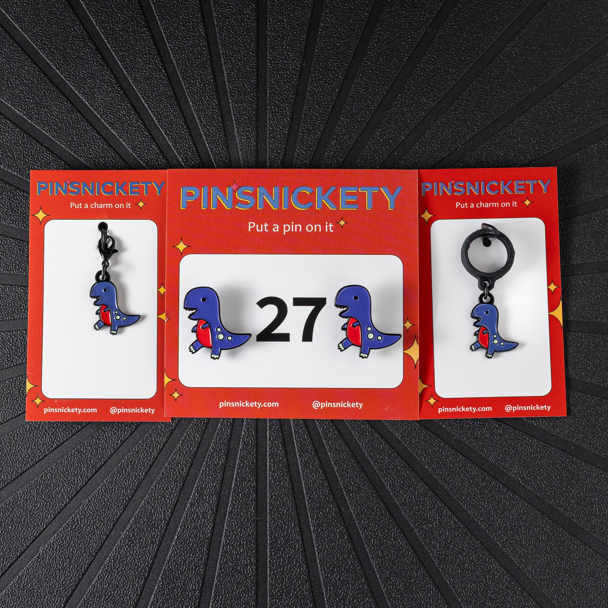 A matching triple combination set with Pinsnickety T-Rex horse show number pins, a bridle charm, and a crop charm in their product packaging on a black background.