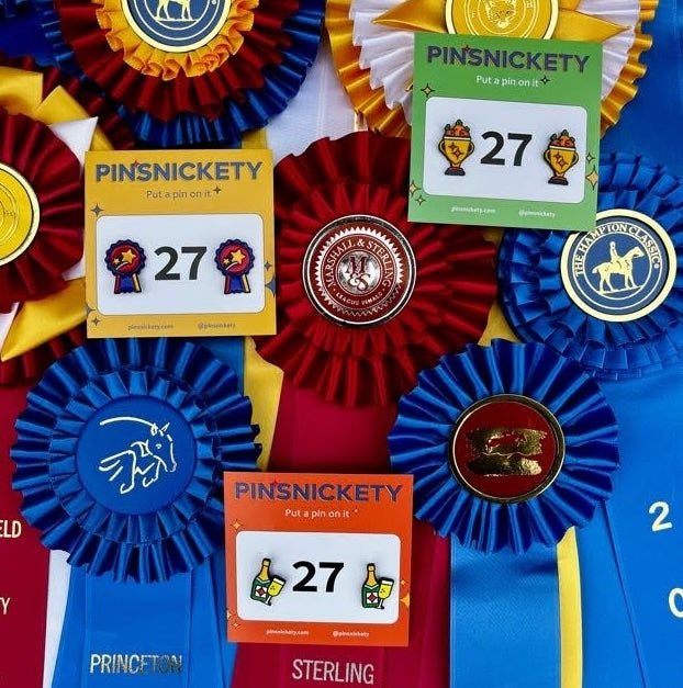pinsnickety champion, champagne and trophy horse show number pins on colorful cards atop a collection of horse show ribbons