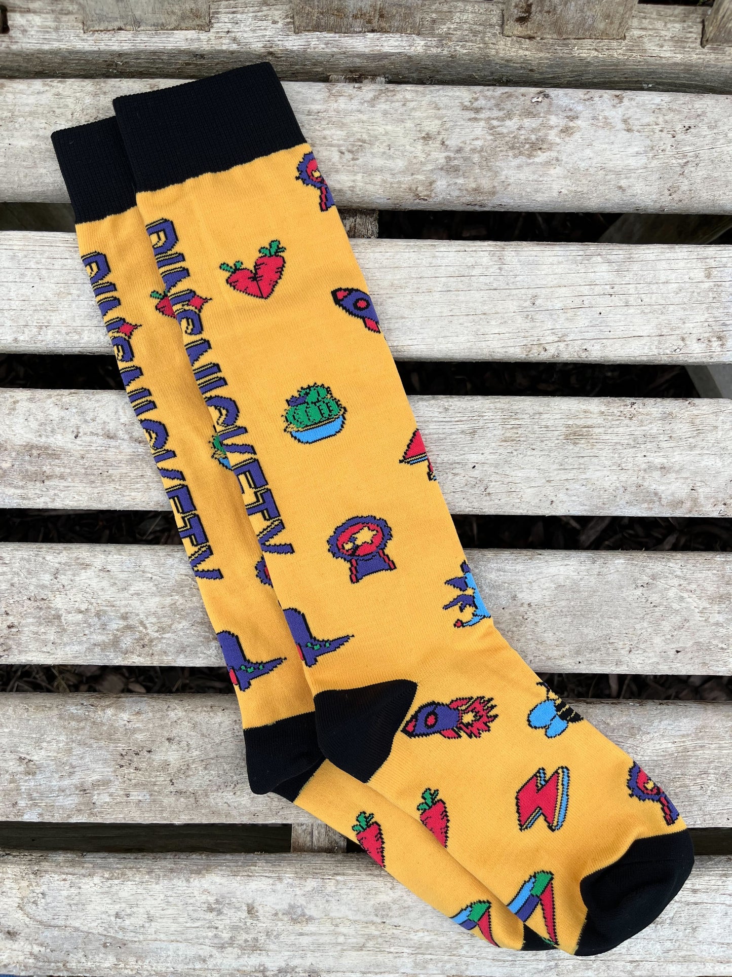 pinsnickety tall boots socks in sunny yellow, covered with pin designs and the company logo, on a wooden slat background